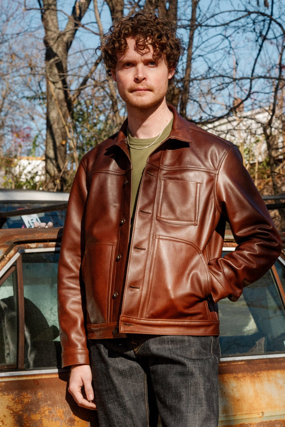 Man in a brown leather jacket standing next to a rusted car in an autumn setting