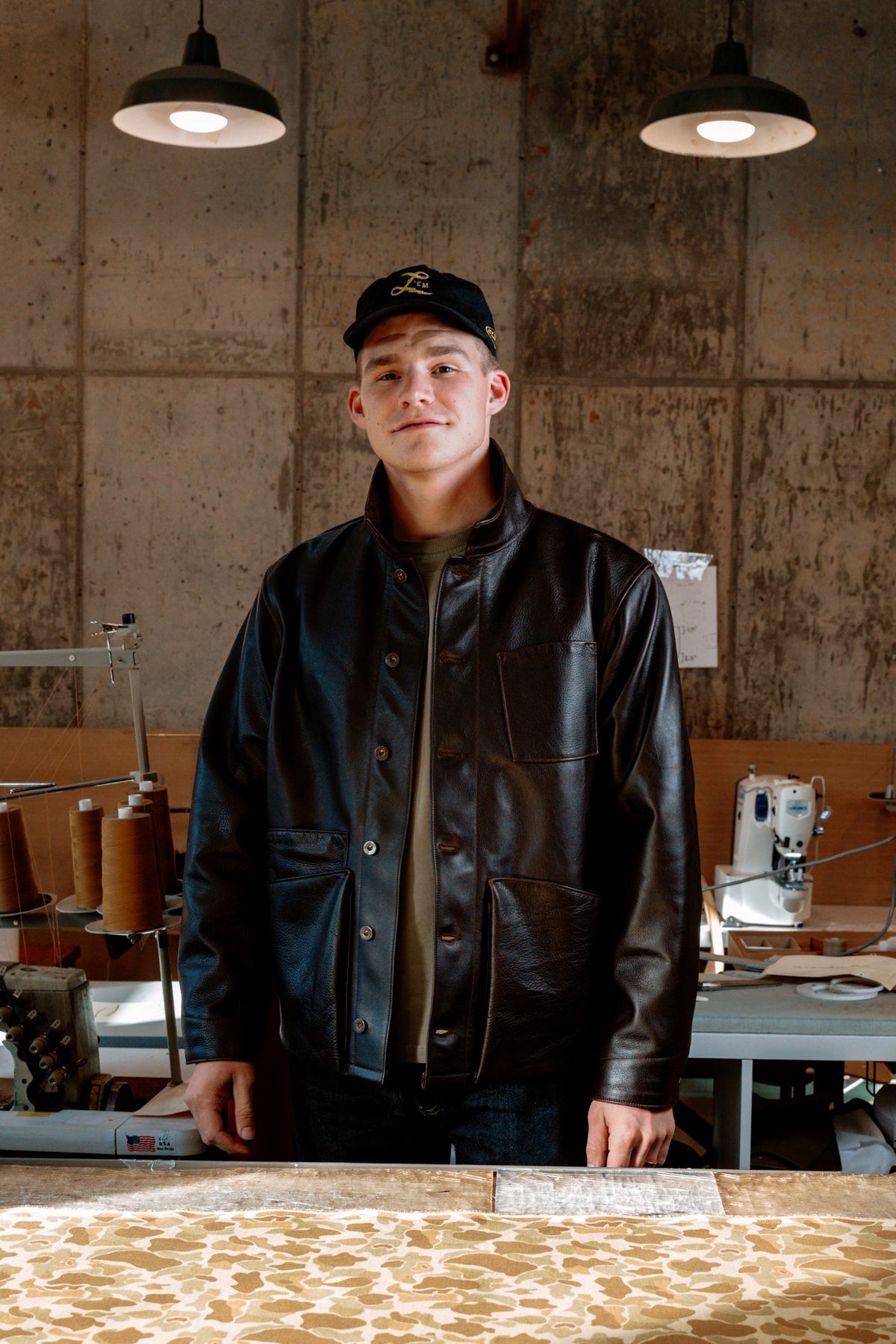 A man wearing a black leather jacket and a black baseball cap stands in a workshop with concrete walls, sewing machines, and a large patterned rug spread out on the floor in the foreground.