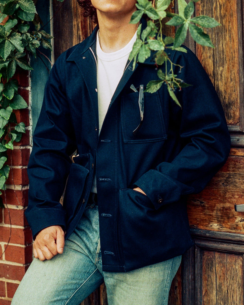 Man leaning against a wooden door with greenery, wearing a dark jacket and light jeans.