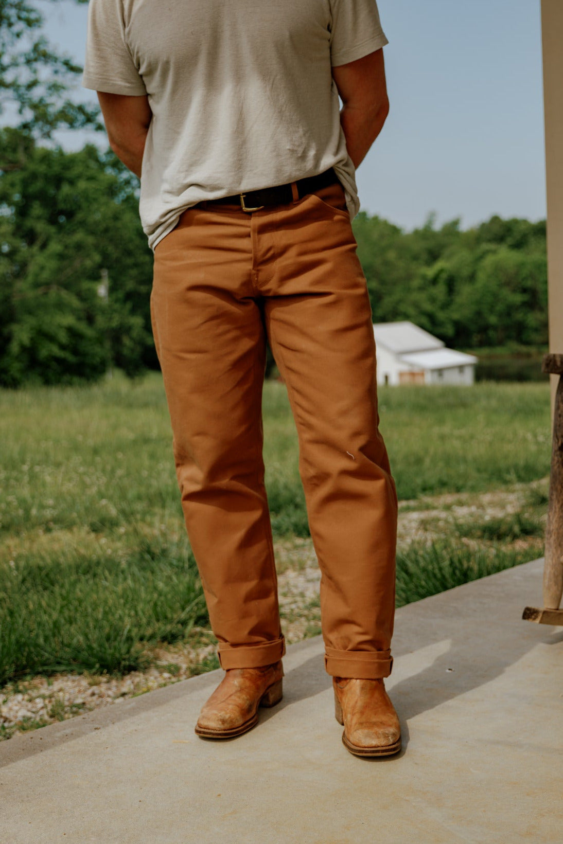 Person wearing a beige t-shirt and brown pants standing on a porch with a green field and trees in the background.