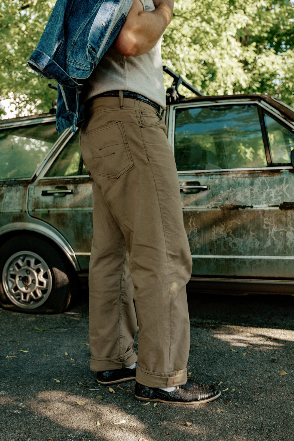 Person wearing beige pants standing next to a vintage car with trees in the background