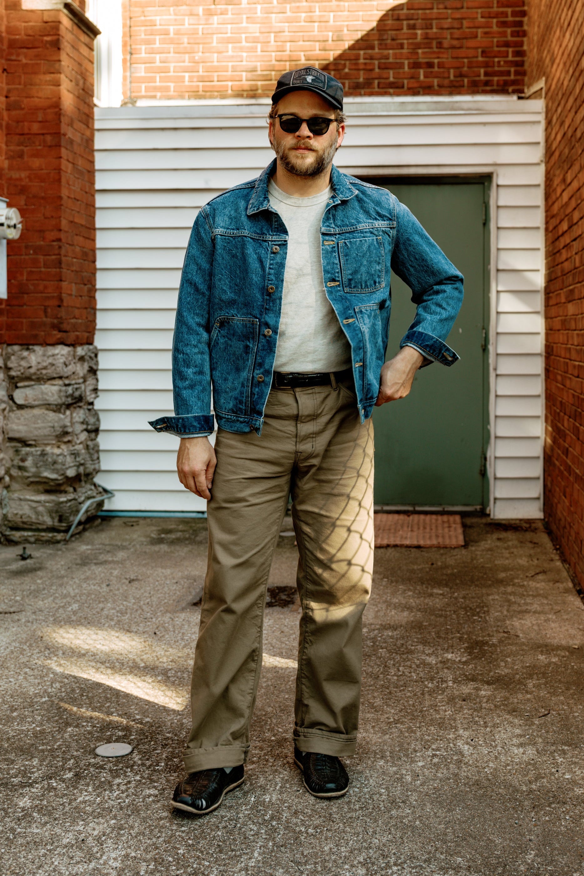 Man wearing a denim jacket and sunglasses standing in an alleyway.