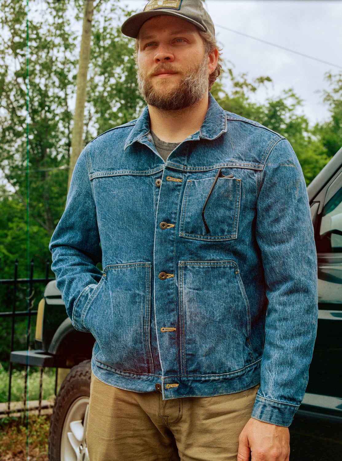 Man wearing a blue denim jacket and cap standing outdoors with trees and a vehicle in the background.