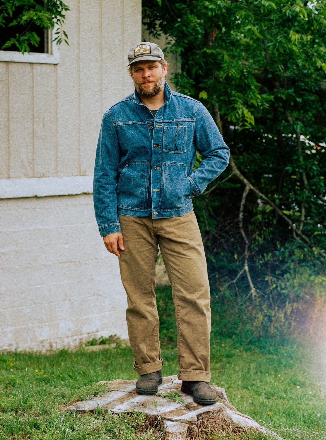 Man wearing a denim jacket and beige pants standing on a stump in front of a white building with greenery.