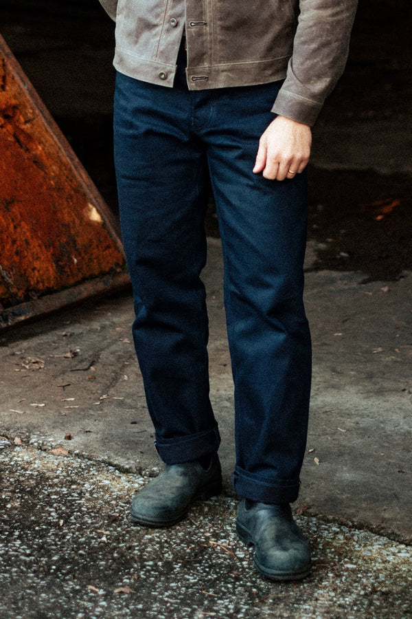 Man wearing a brown jacket and blue jeans standing in a dark, industrial setting.